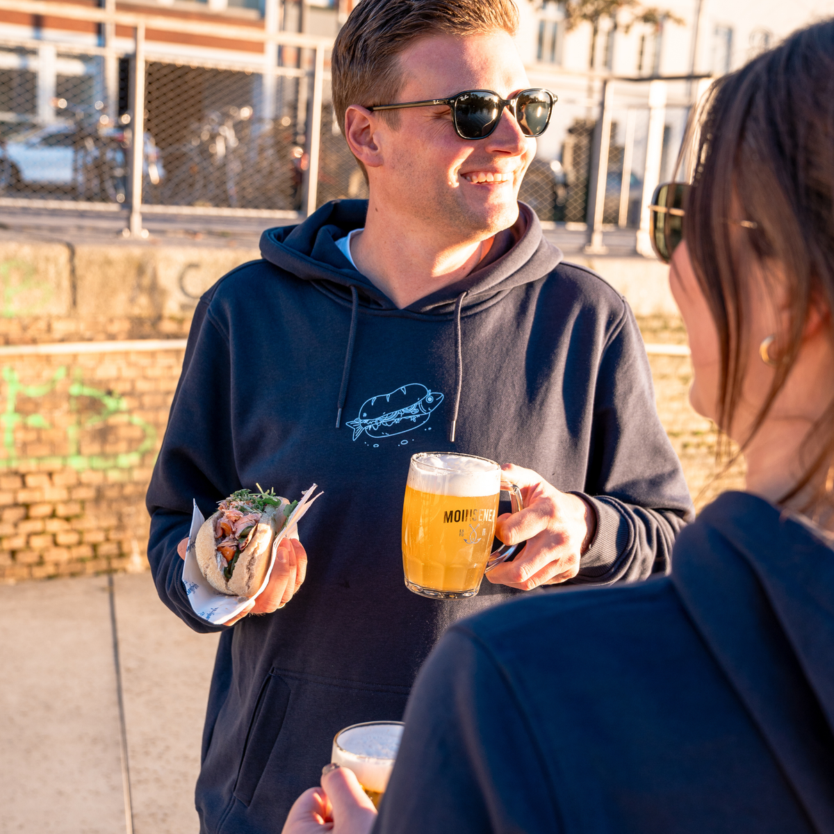 Mann lächelt mit Sonnenbrille und hält ein Fischbrötchen & ein Bier