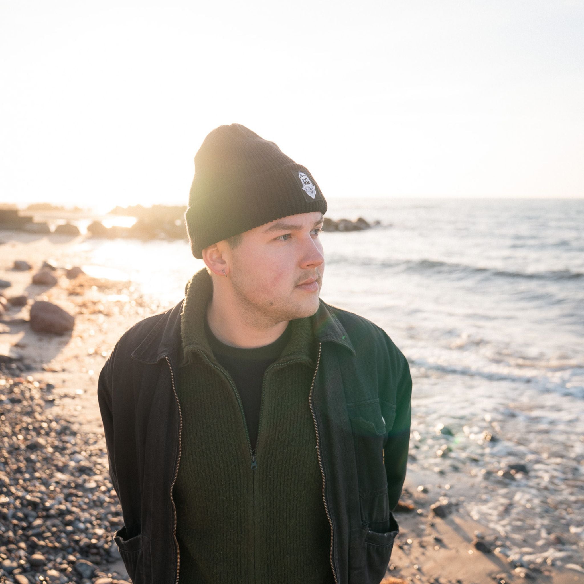 Junger Mann am Strand mit Matrosenmütze, schwarze Wolle, vor einer sonnigen Küstenlandschaft.