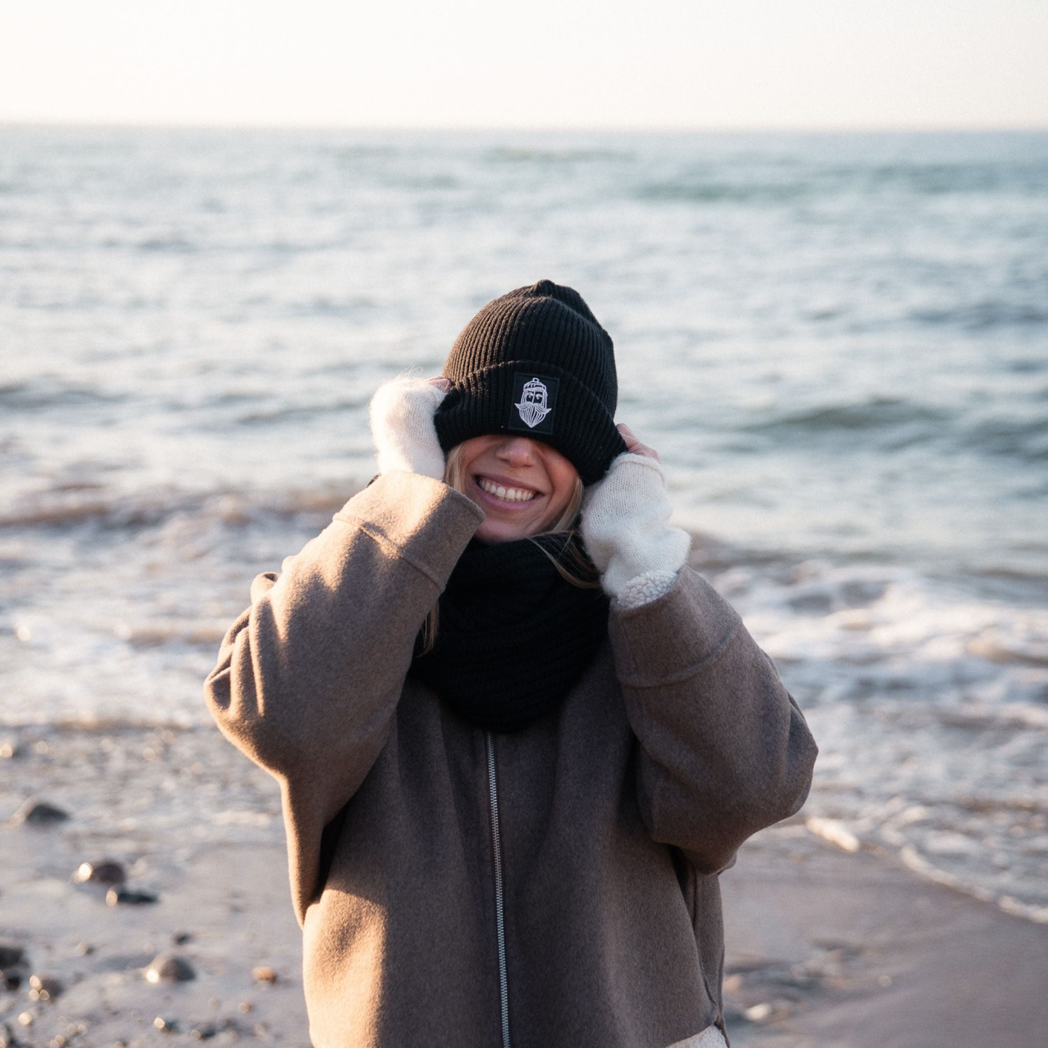Junge Frau am Strand mit Matrosenmütze und wolliger Jacke, lächelt und genießt die kalte Meeresluft.
