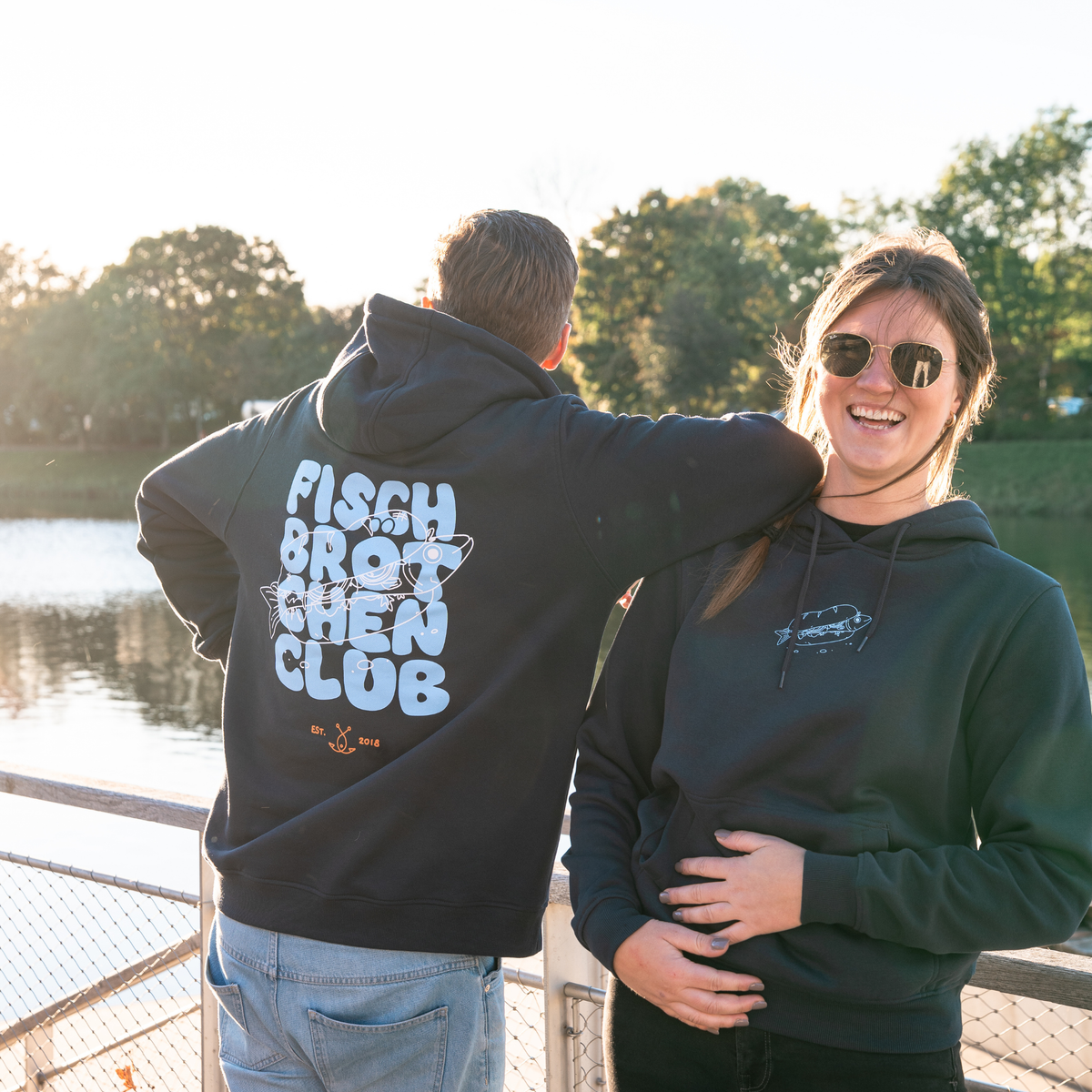 Paar steht am Waser mit Fischbrötchenclub Hoodies, sie schaut in die Kamera, er steht mit dem Rücken zu uns