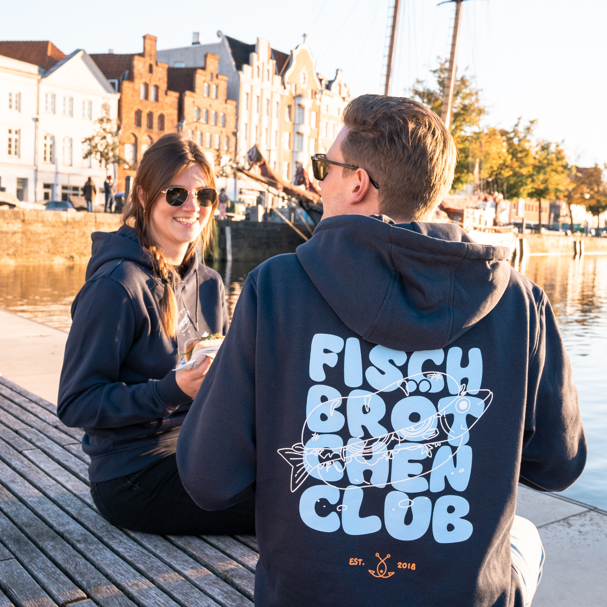 Paar sitzt am Wasser, er mit dem Rücken zu uns und einem großen Schriftzug auf seinem Hoodie