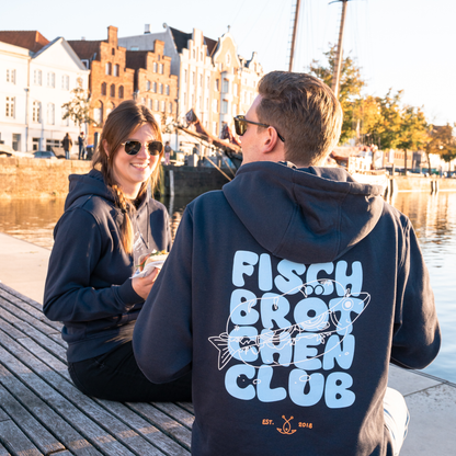Paar sitzt am Wasser, er mit dem Rücken zu uns und einem großen Schriftzug auf seinem Hoodie