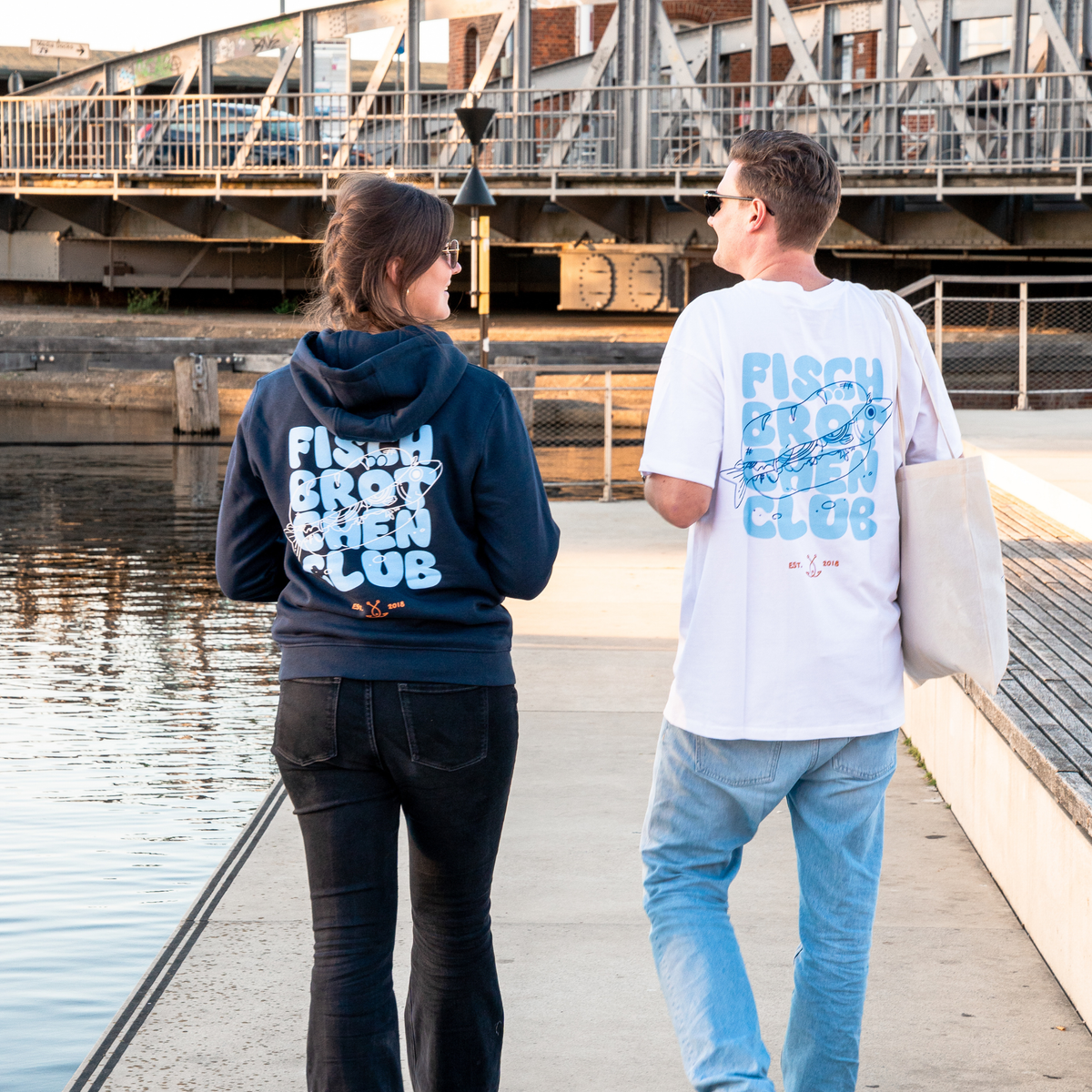 Paar läuft auf eine Brücke zu, sie hat einen dunkeln Hoodie mit einem großen Schriftzug an und er ein weißes Shirt, mit dem selben Schriftzug