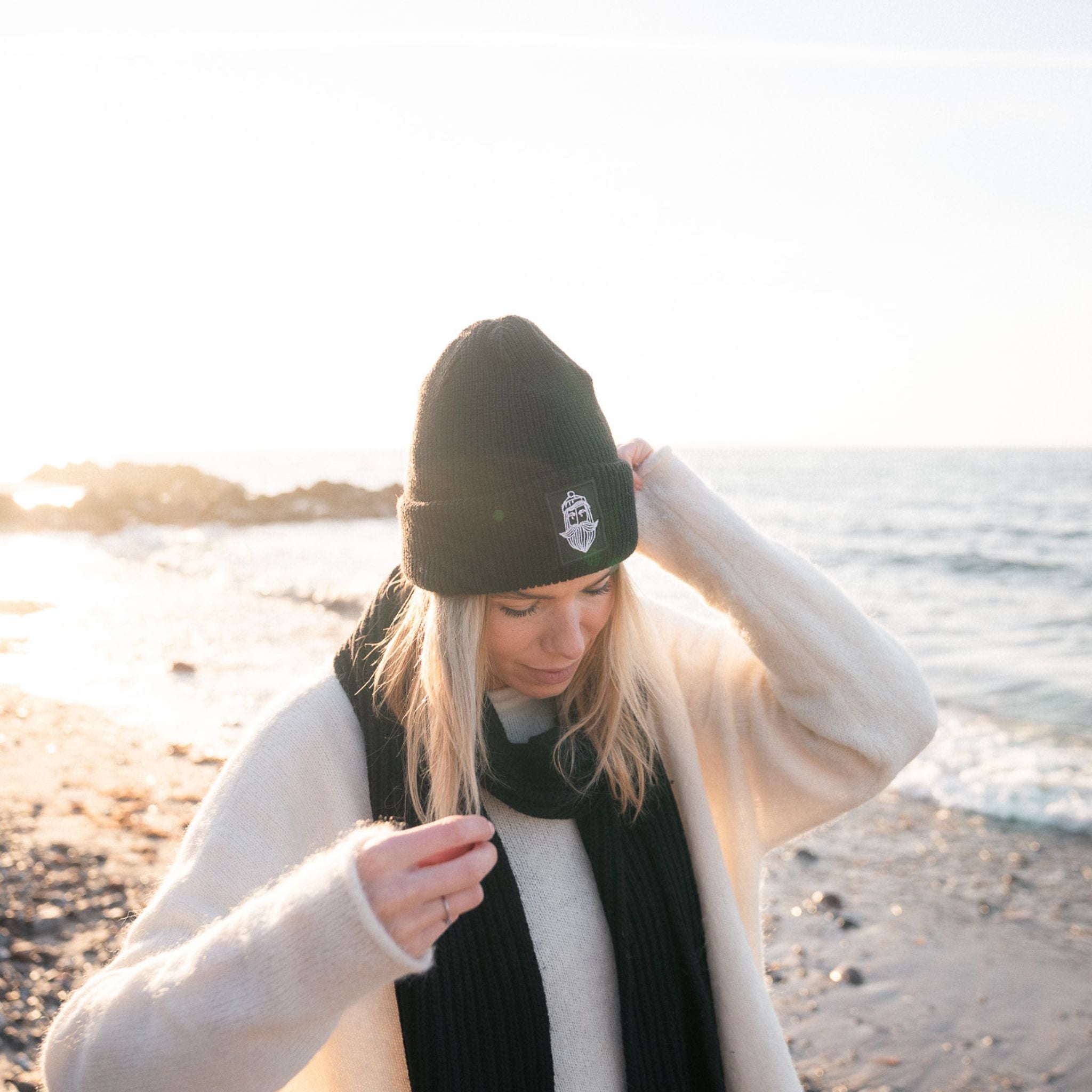 Frau am Strand mit Matrosenmütze und schwarzem Schal, Sonne und Wasser im Hintergrund.