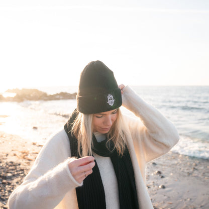 Frau am Strand mit Matrosenmütze und schwarzem Schal, Sonne und Wasser im Hintergrund.