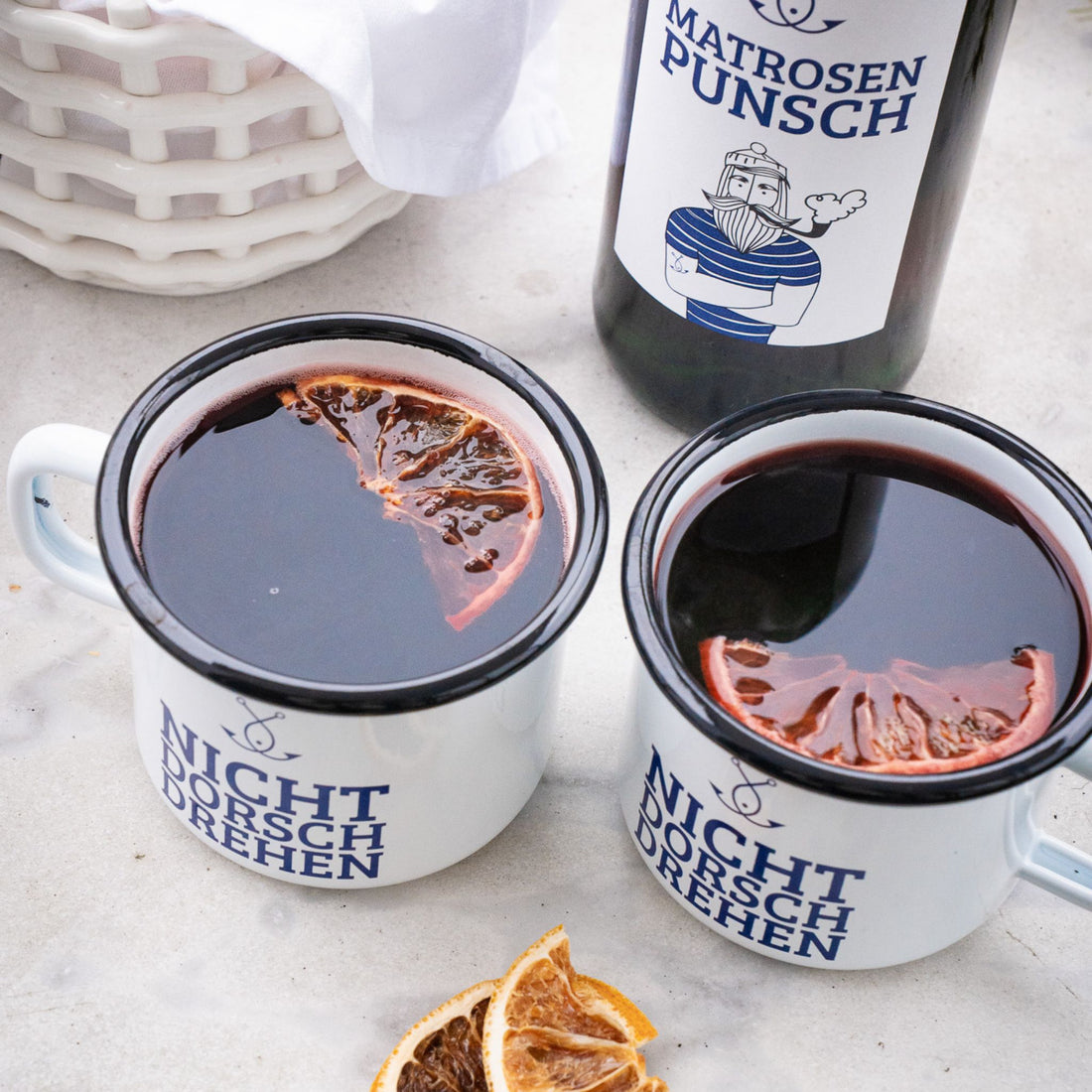 Zwei Emaille-Tassen mit Glühwein und Matrosenpunsch Flasche im Hintergrund auf hellem Tisch