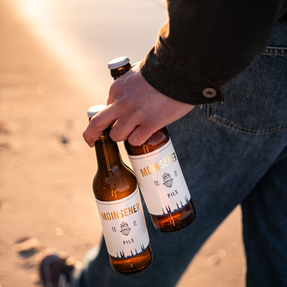 2 Moinsener Pils Bierflaschen, die von einem Mann in der Hand getragen werden