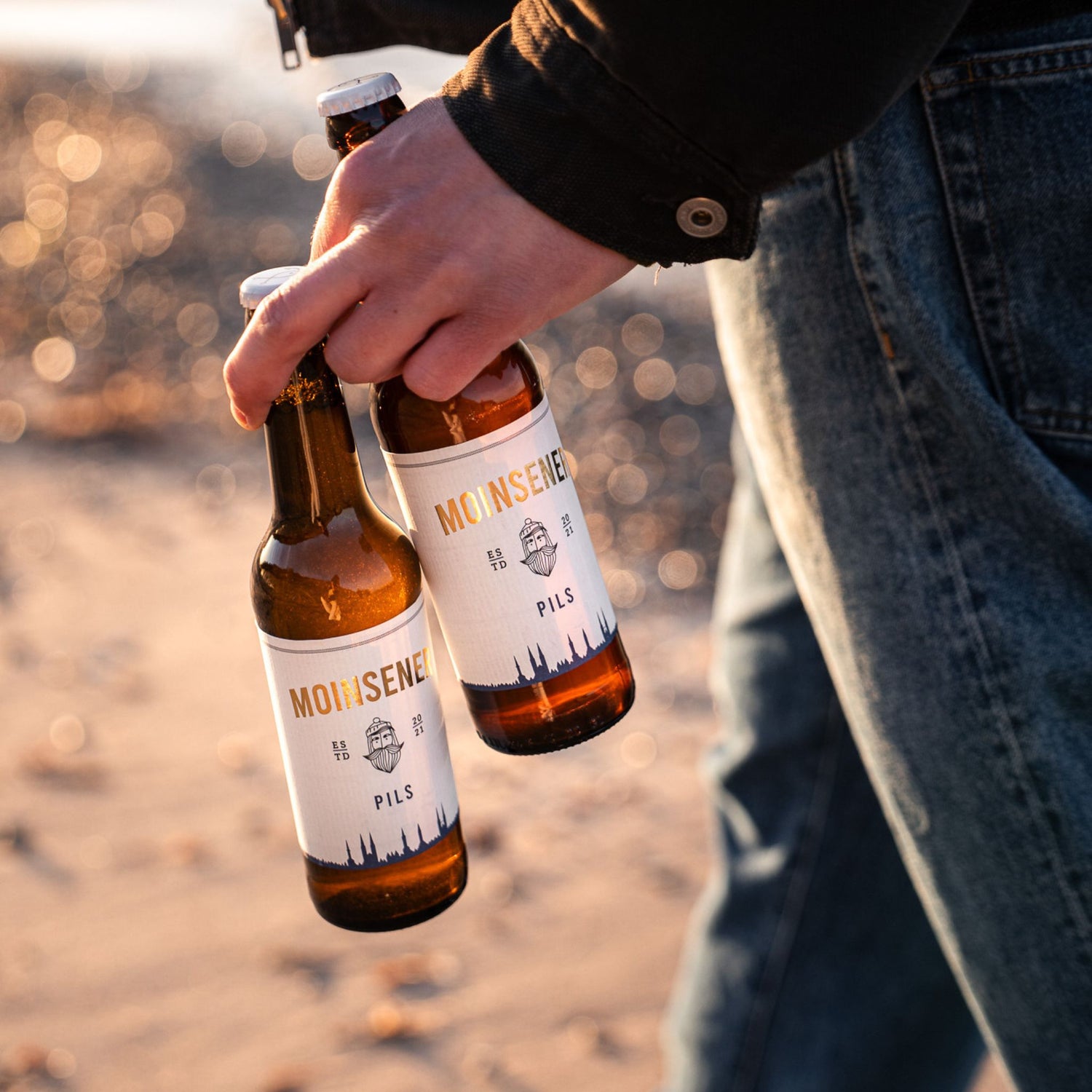 2 Moinsener Pils Bierflaschen, die von einem Mann in der Hand getragen werden