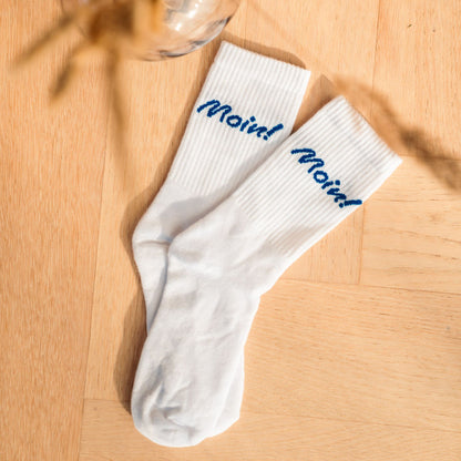 Weiße Fangfrisch Socken mit blauem „Moin“ Schriftzug auf hellem Holzboden